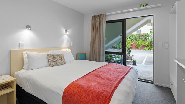 A hotel room with a bed, a nightstand, a window with curtains, a door with an exit sign, and a view of a staircase and greenery outside.