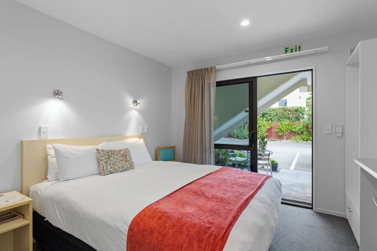 Une chambre d’hôtel avec un lit, une table de chevet, une fenêtre avec des rideaux, une porte avec un panneau indiquant la sortie, et une vue sur un escalier et de la verdure à l’extérieur.