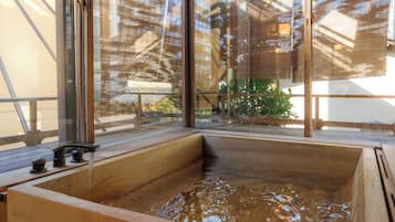 A wooden hot tub with a view of a building through a window with bamboo blinds.