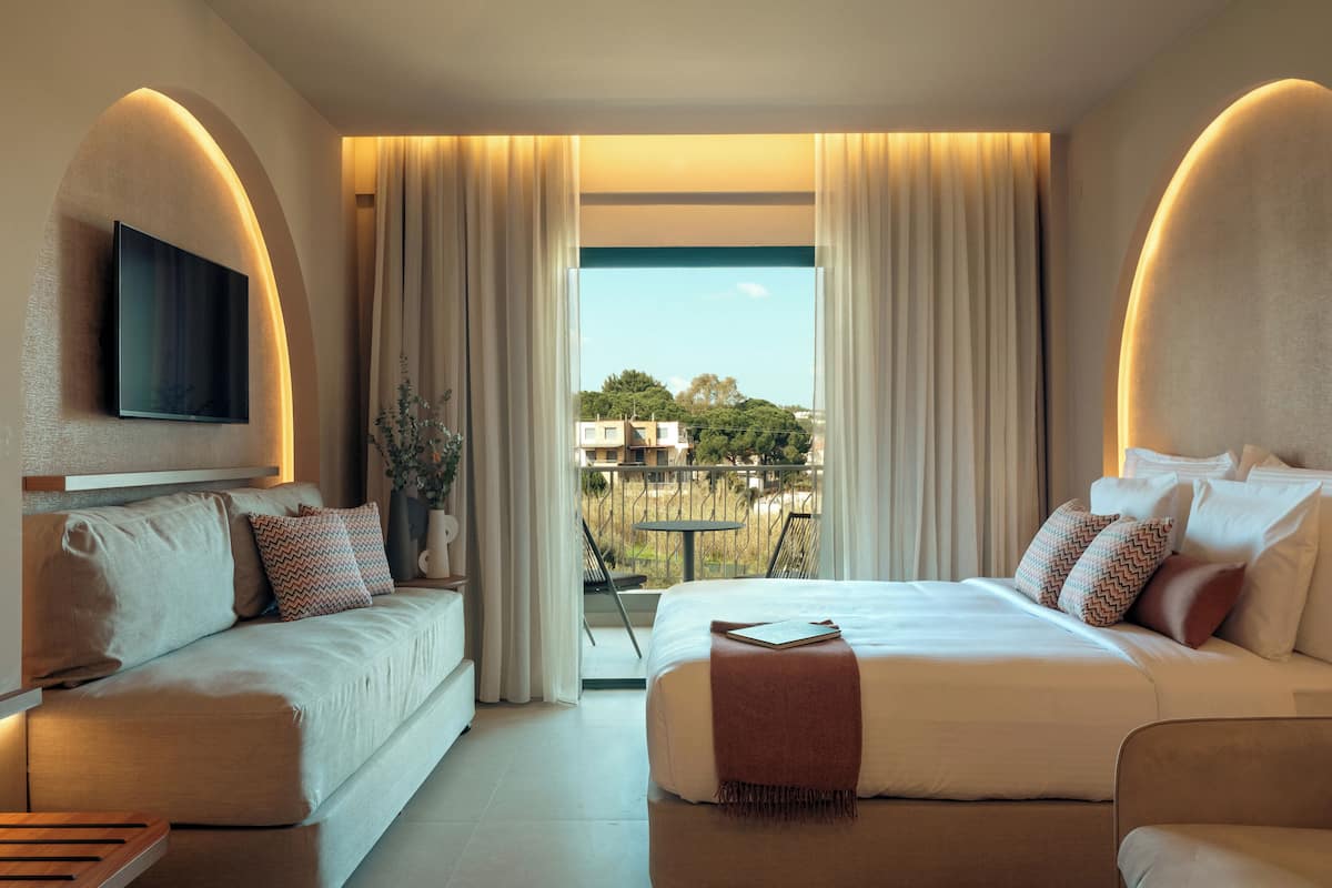 Une chambre d’hôtel avec un lit, un canapé, un téléviseur fixé au mur et un balcon offrant une vue sur les bâtiments et les arbres.