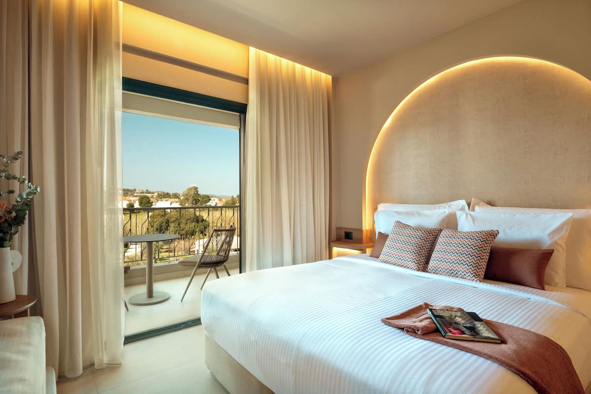 Une chambre d’hôtel avec un grand lit, une vue sur l’extérieur, un balcon avec une chaise et un luminaire rond au-dessus du lit.