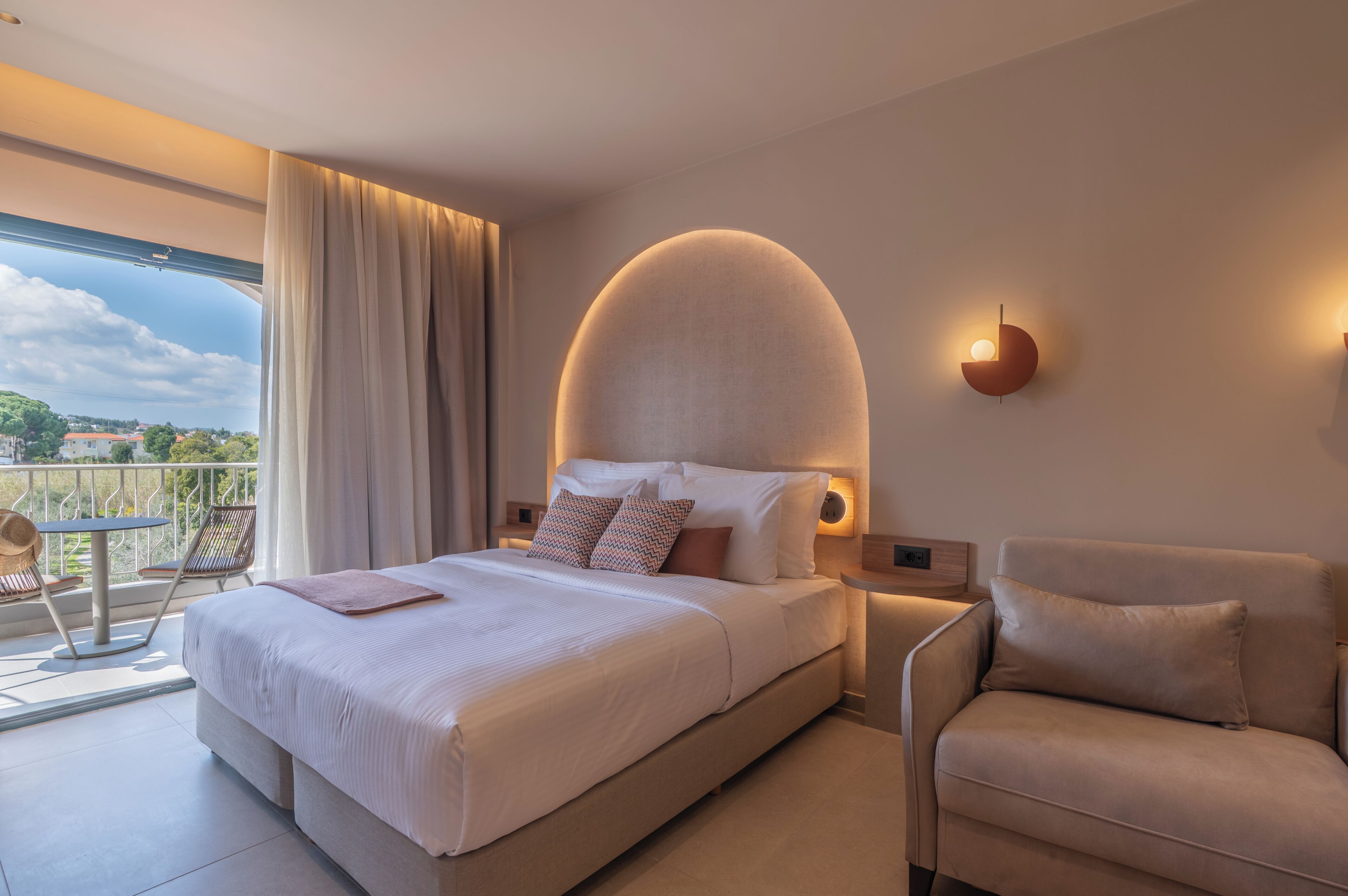 A modern hotel room with a large bed, a sofa, a balcony with a view, and a unique ceiling light fixture.