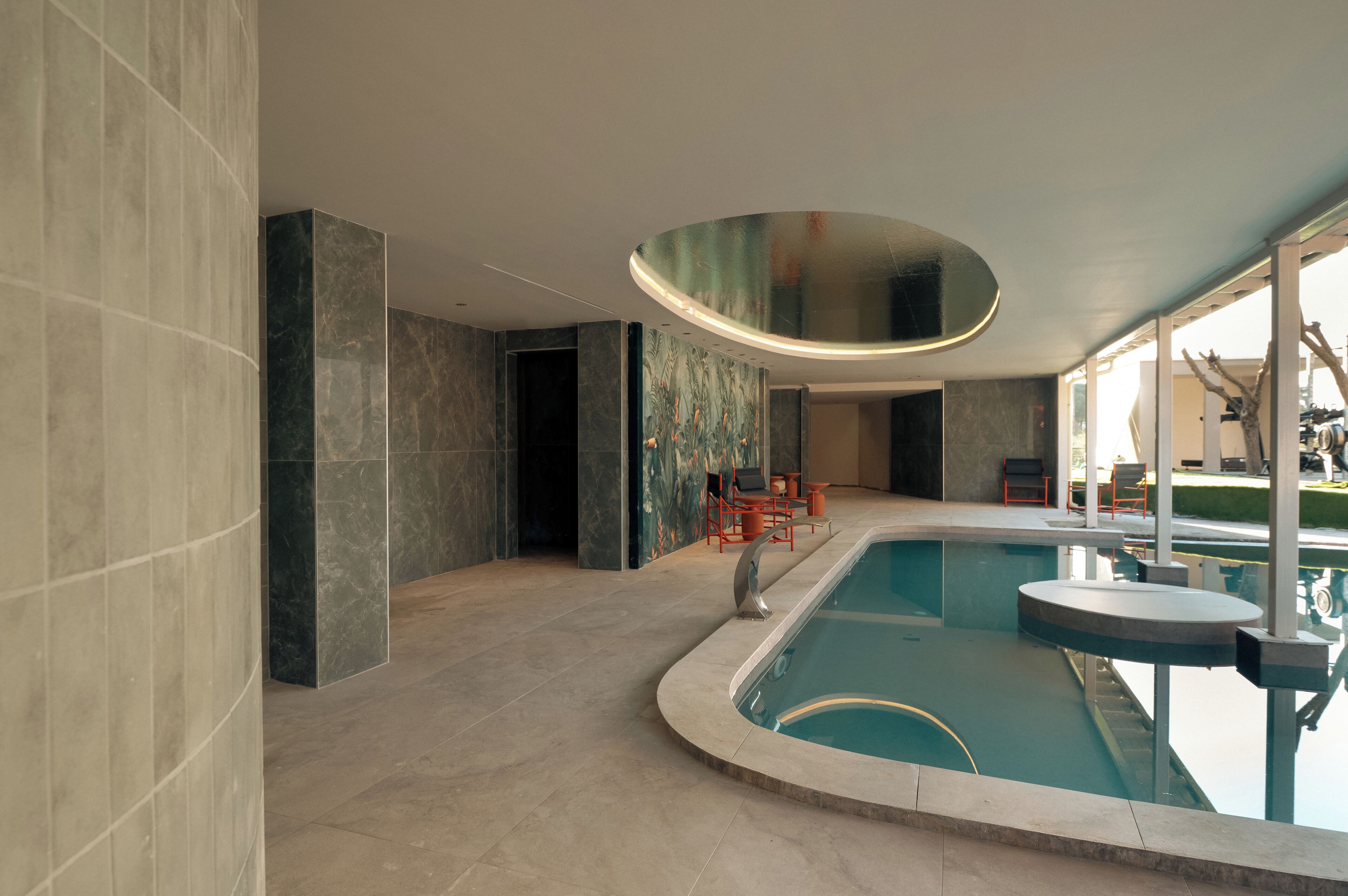 A modern indoor swimming pool with a curved ceiling, stone walls, and a seating area with chairs and a table.