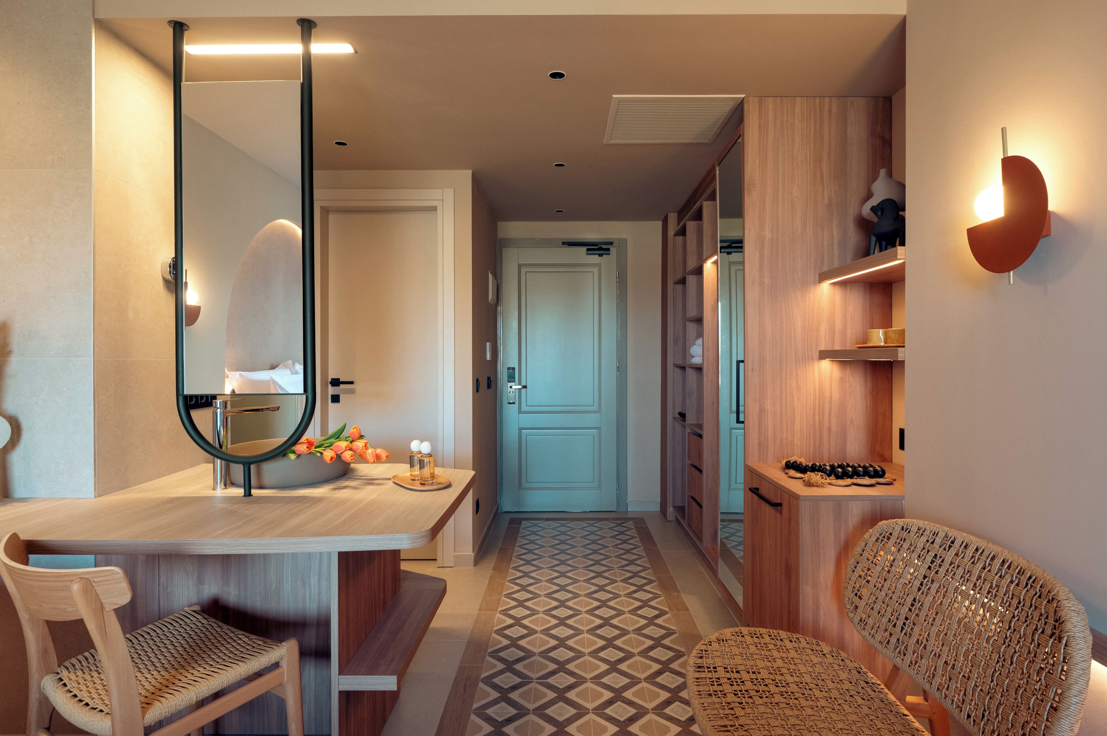 Un couloir d’hôtel moderne avec un comptoir d’accueil en bois, un miroir, un banc, des chaises en osier, un tapis à motifs et une porte bleue.