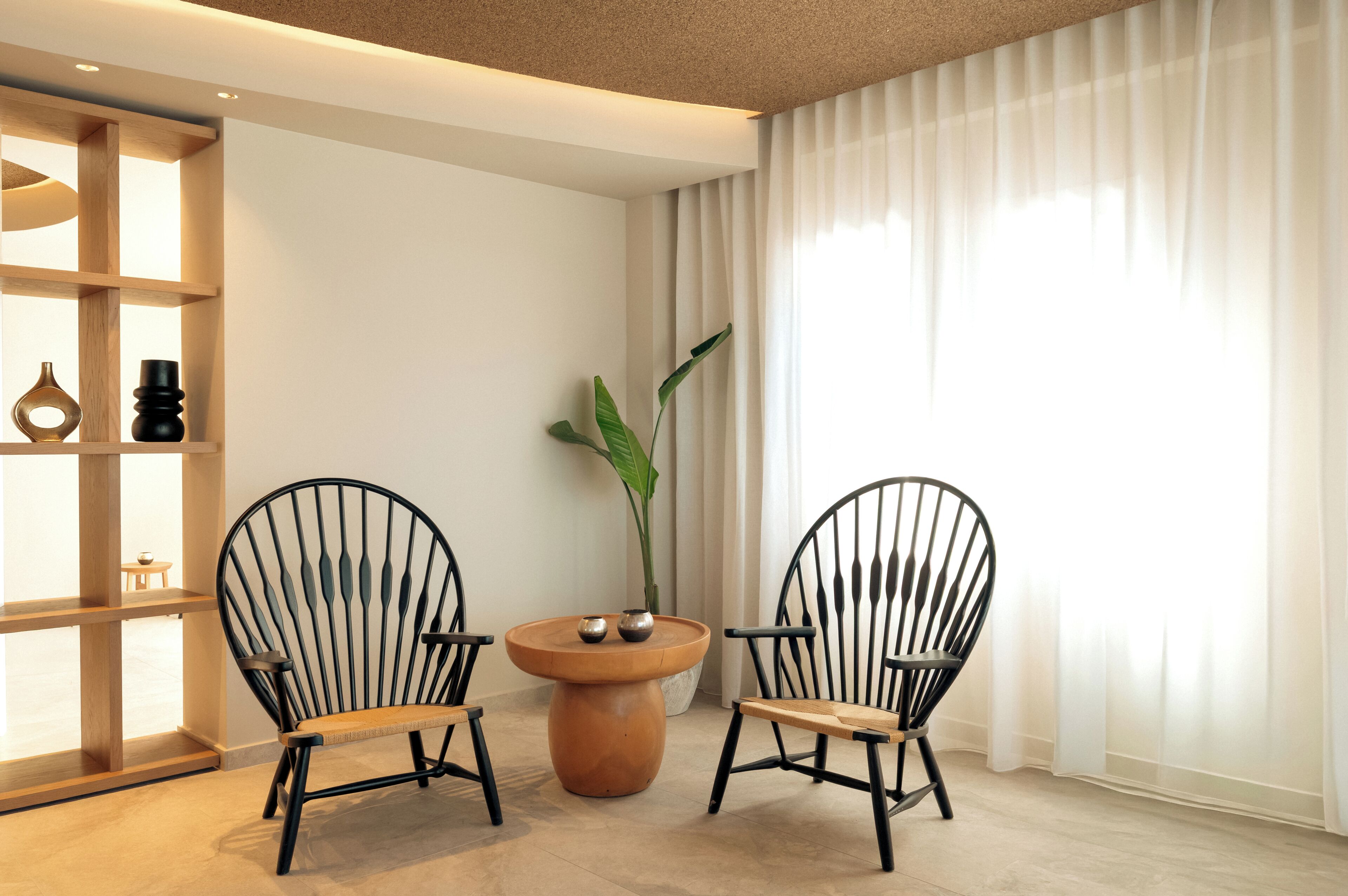 Une pièce moderne avec deux chaises noires, une table ronde en bois, une plante en pot et une étagère en bois adossée à un mur blanc.
