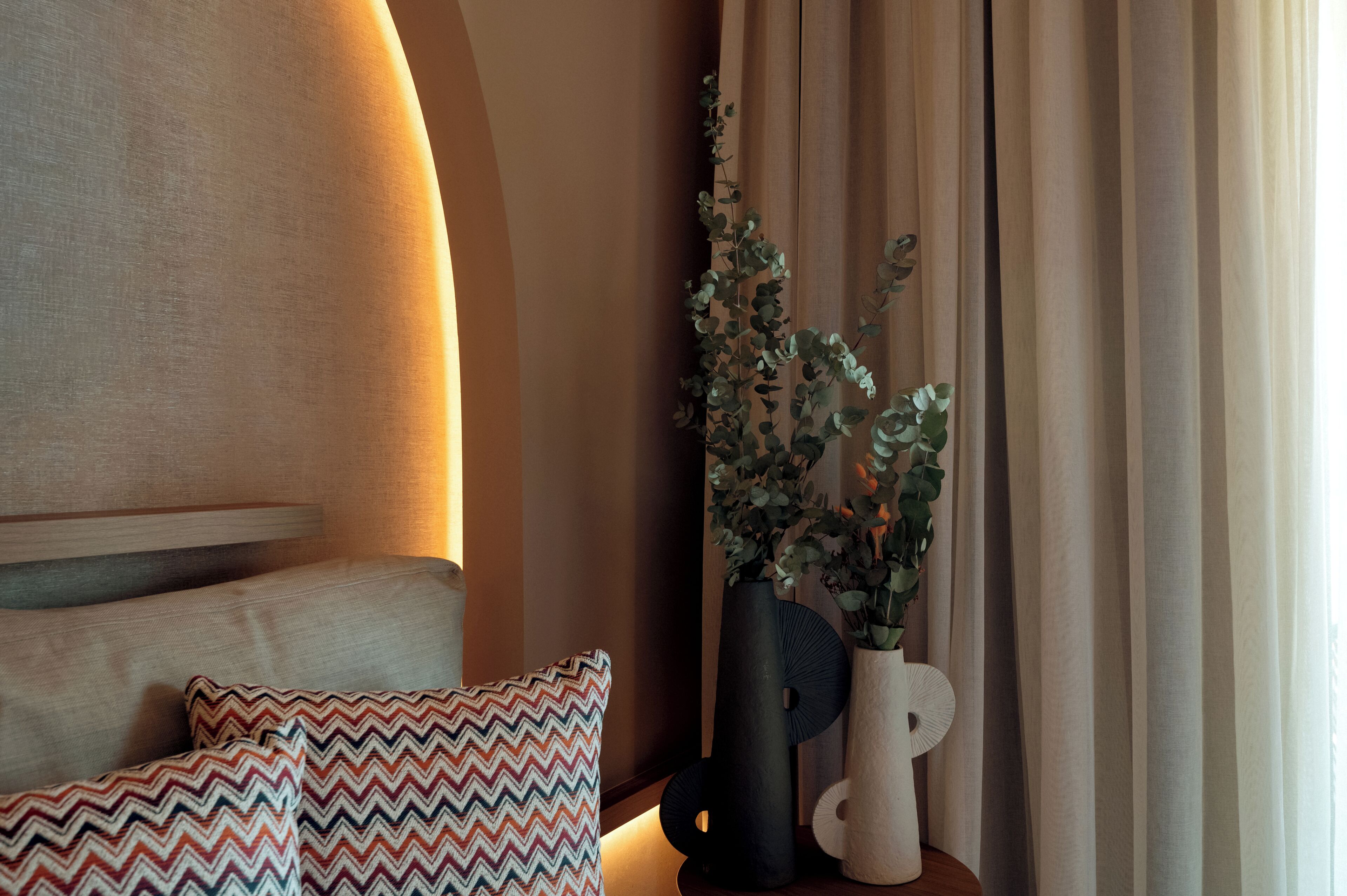 A room with a bed, pillows, a vase with plants, and curtains.