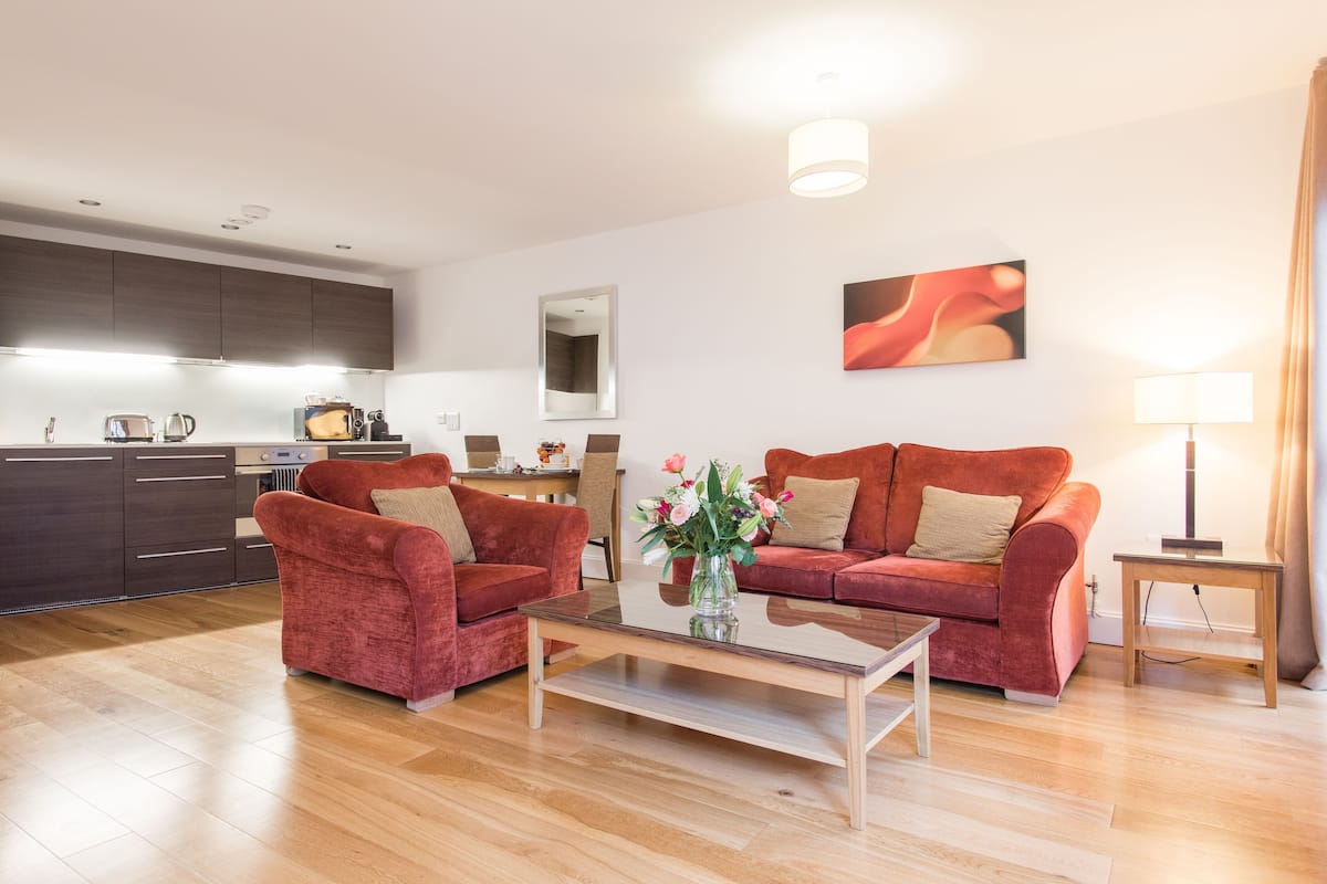 Un salon moderne avec une table basse en verre, deux canapés rouges, une cuisine en arrière-plan et un tableau au mur.