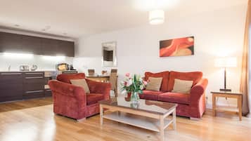 A modern living room with a glass coffee table, two red sofas, a kitchen in the background, and a painting on the wall.