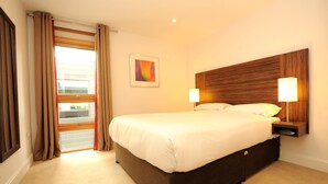 A hotel room with a large bed, a wooden headboard, two bedside lamps, a window with curtains, and a framed picture on the wall.