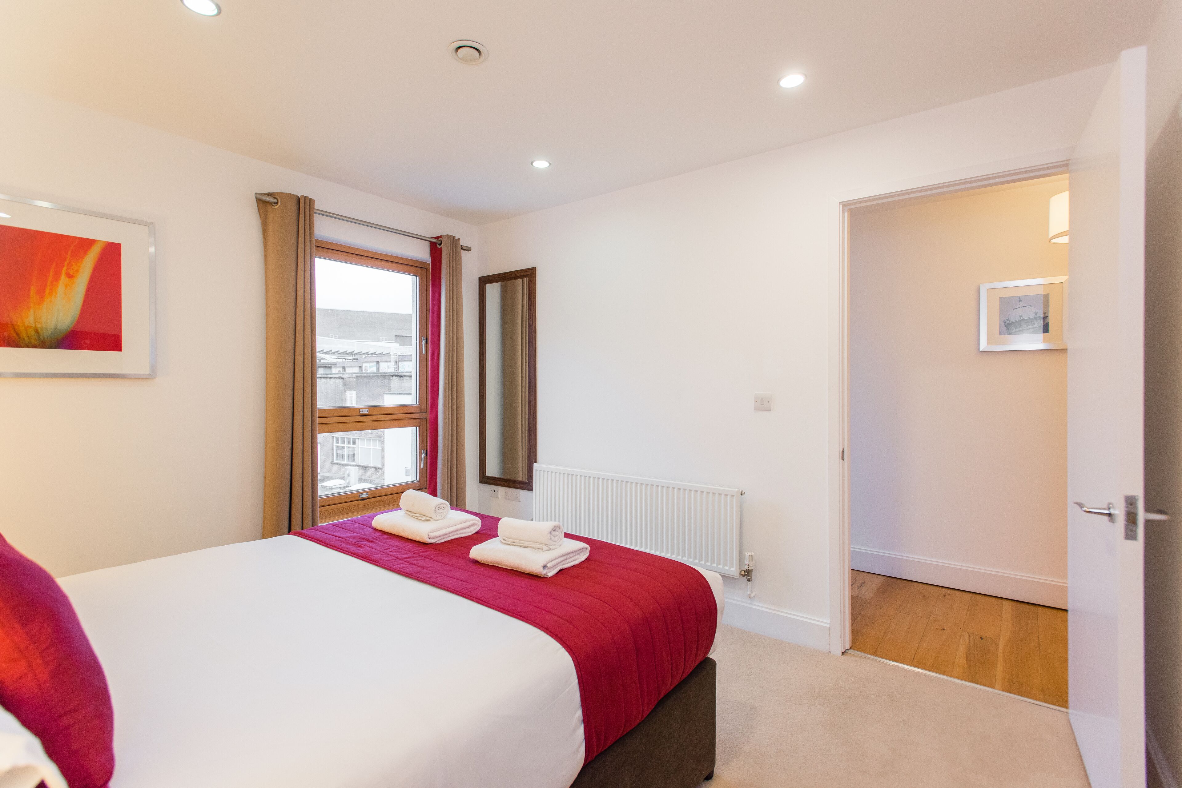 Un dormitorio con cama, ventana con cortinas, radiador y puerta que da a otra habitación.