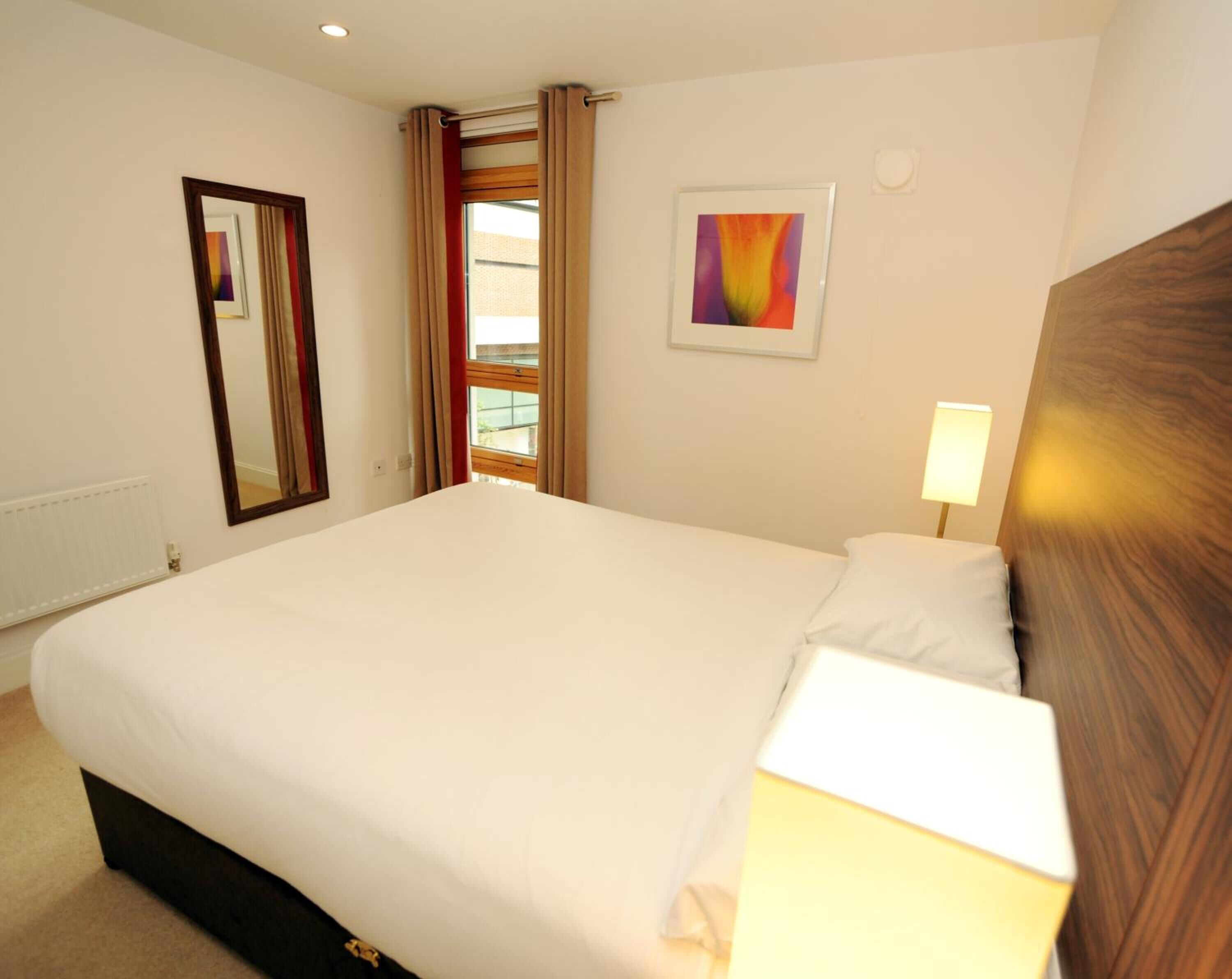 A hotel room with a large bed, a bedside lamp, a window with curtains, a framed picture on the wall, and a mirror.
