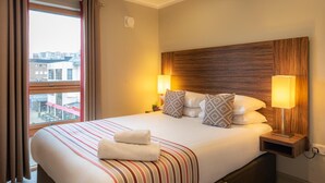 A hotel room with a large bed, two bedside lamps, a window with curtains, and a view of buildings outside.