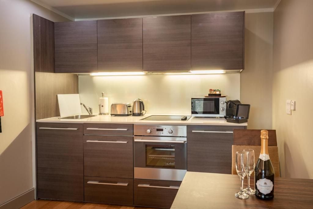 A modern kitchen with dark cabinetry, a built-in oven, a microwave, and a dining area with a bottle of champagne.