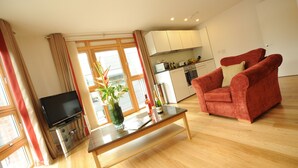 A living room with a TV, a glass coffee table, a red armchair, and a kitchen in the background.