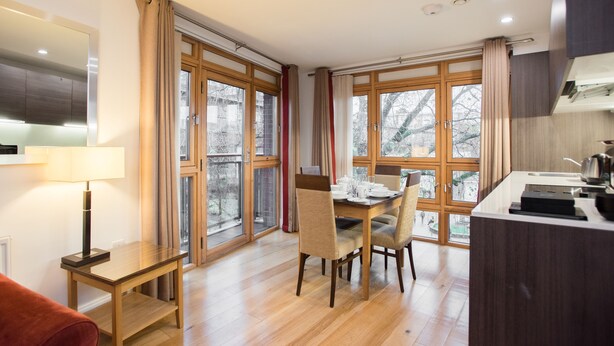 A modern kitchen with wooden cabinets, a dining table with chairs, and large windows with curtains.