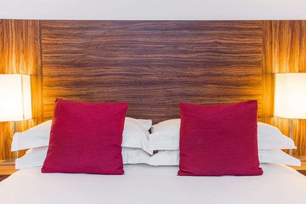 A bed with white linens and two red pillows, flanked by two bedside lamps with white shades, against a wooden headboard.