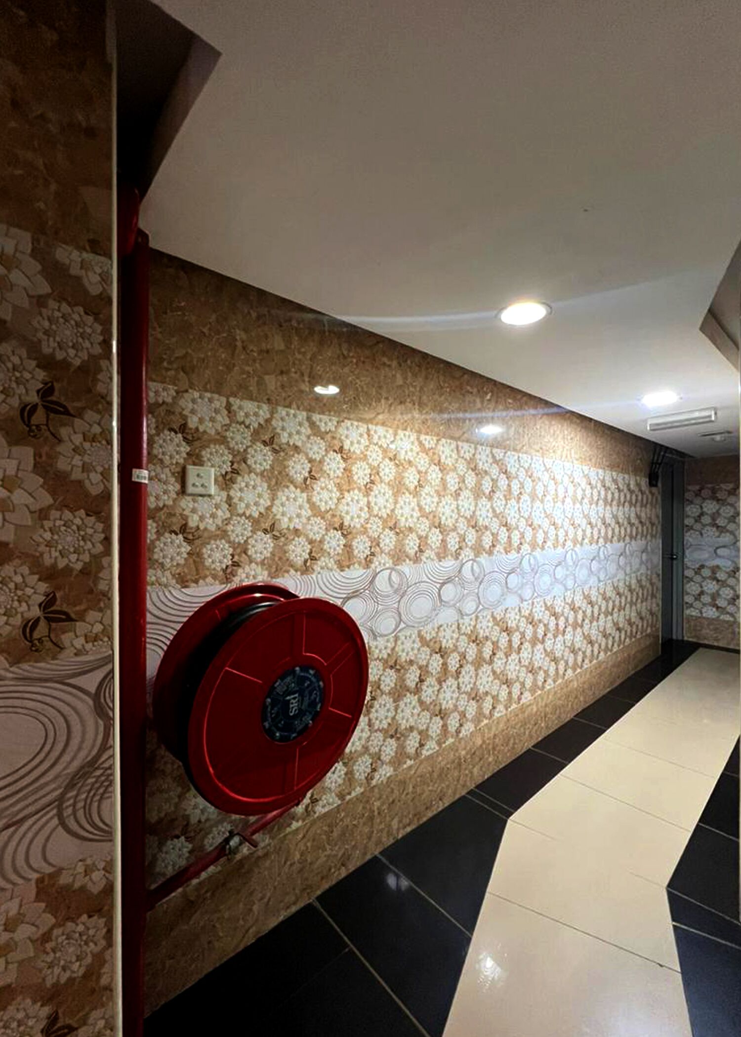 Um corredor com ladrilhos estampados na parede, um alarme de incêndio vermelho e piso ladrilhado.