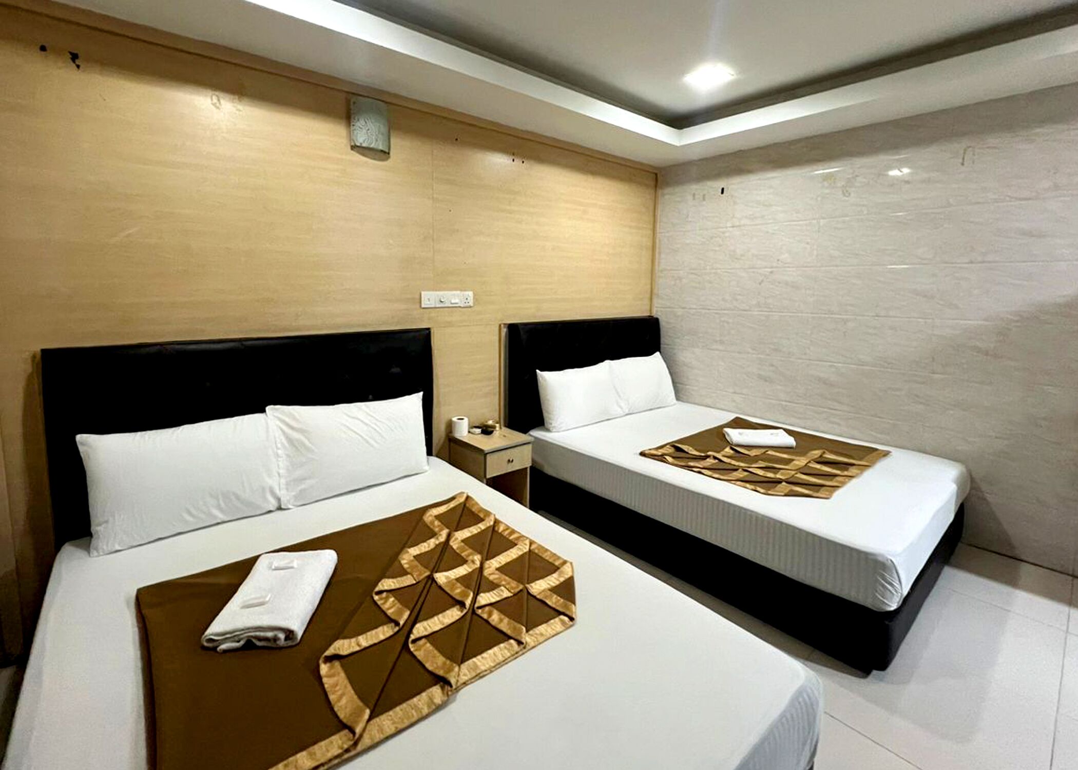 Two single beds with white linens, a wooden headboard, a nightstand with a lamp, and a wall-mounted light fixture.