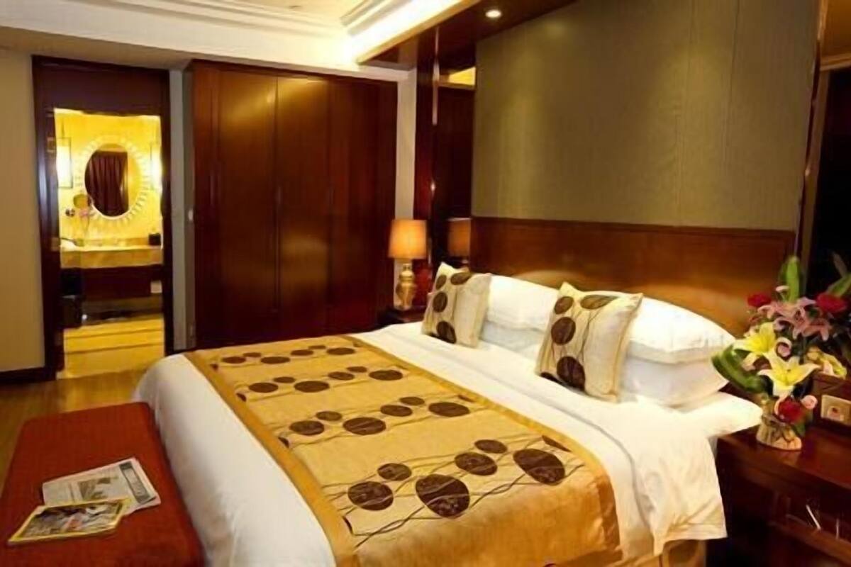 A hotel room with a large bed, a wooden wardrobe, a mirror, a nightstand with a lamp, and a floral arrangement.