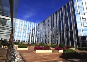 Um edifício moderno com fachada de vidro refletante, deck de madeira e canteiros de flores ajardinados.