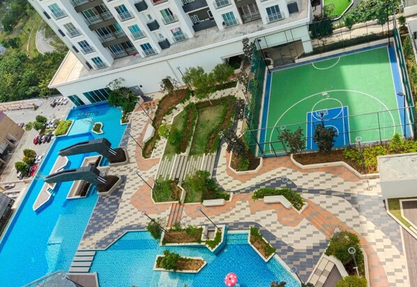 A rooftop pool area with a basketball court, surrounded by a checkered tile pattern and greenery.