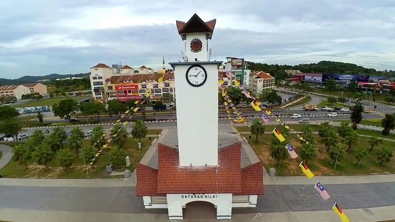 A clock tower with a white base and a red roof, surrounded by palm trees and buildings.