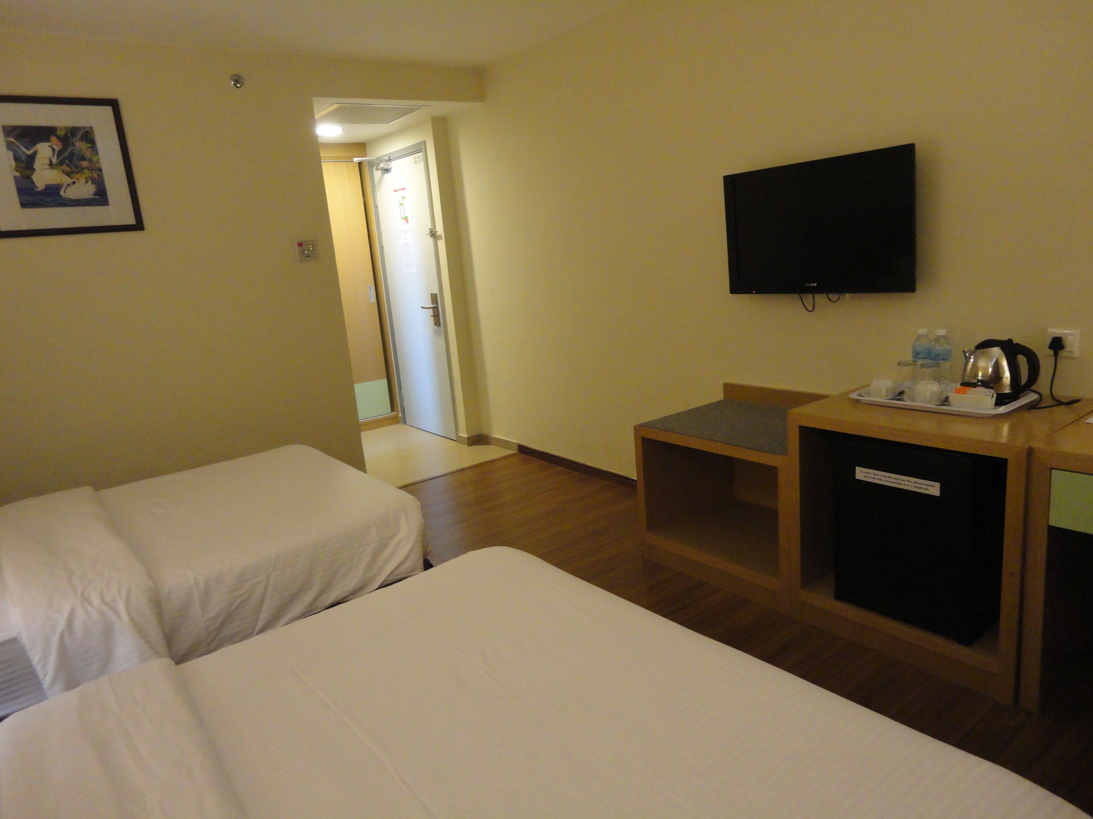 A hotel room with two beds, a desk with a kettle and cups, a flat-screen TV, and a framed picture on the wall.