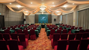 Un salón de conferencias con filas de mesas y sillas, un escenario con un atril y una pantalla grande mostrando un logotipo.
