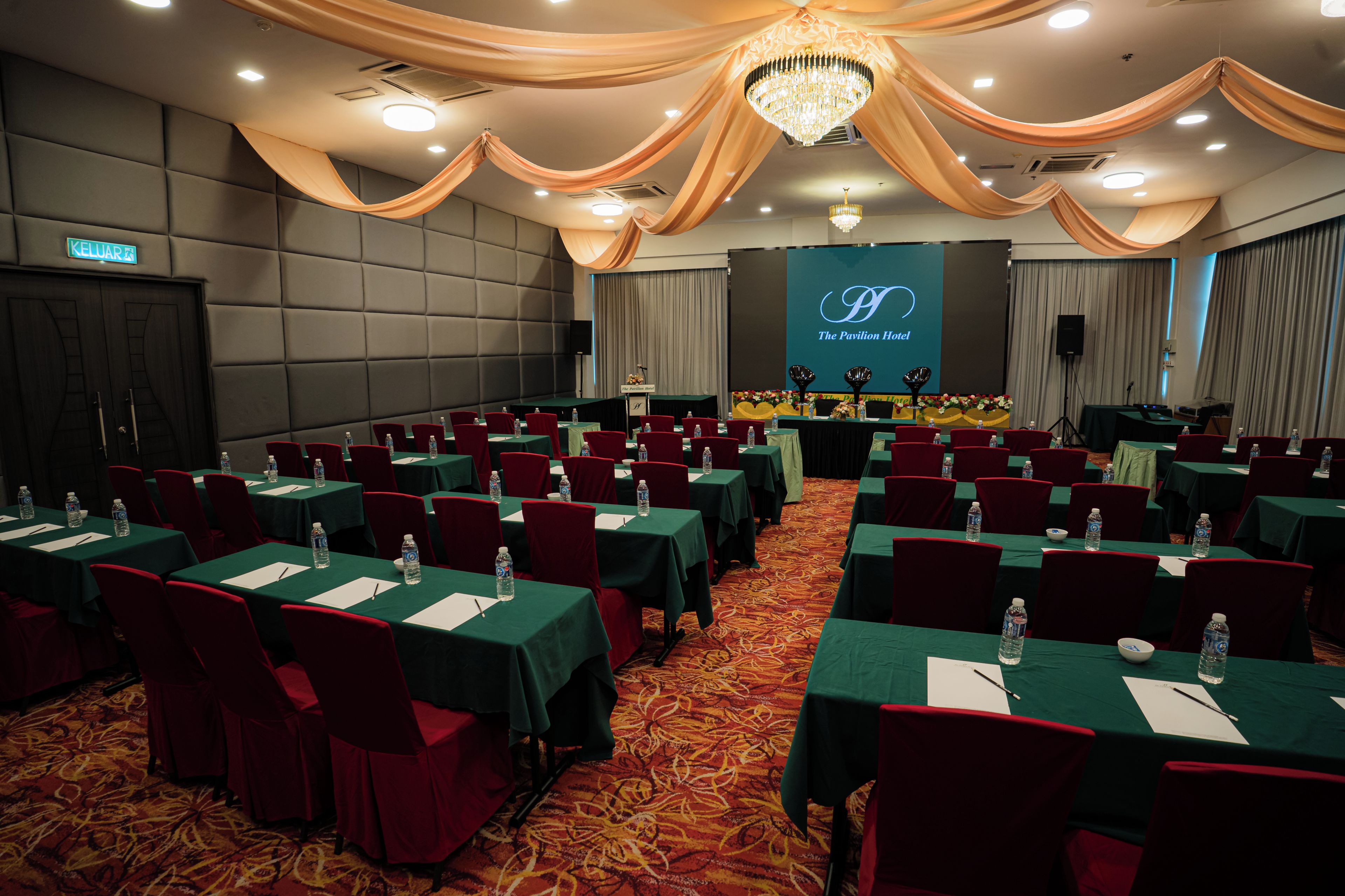 A conference room with rows of tables and chairs, a stage with a screen, and a chandelier.