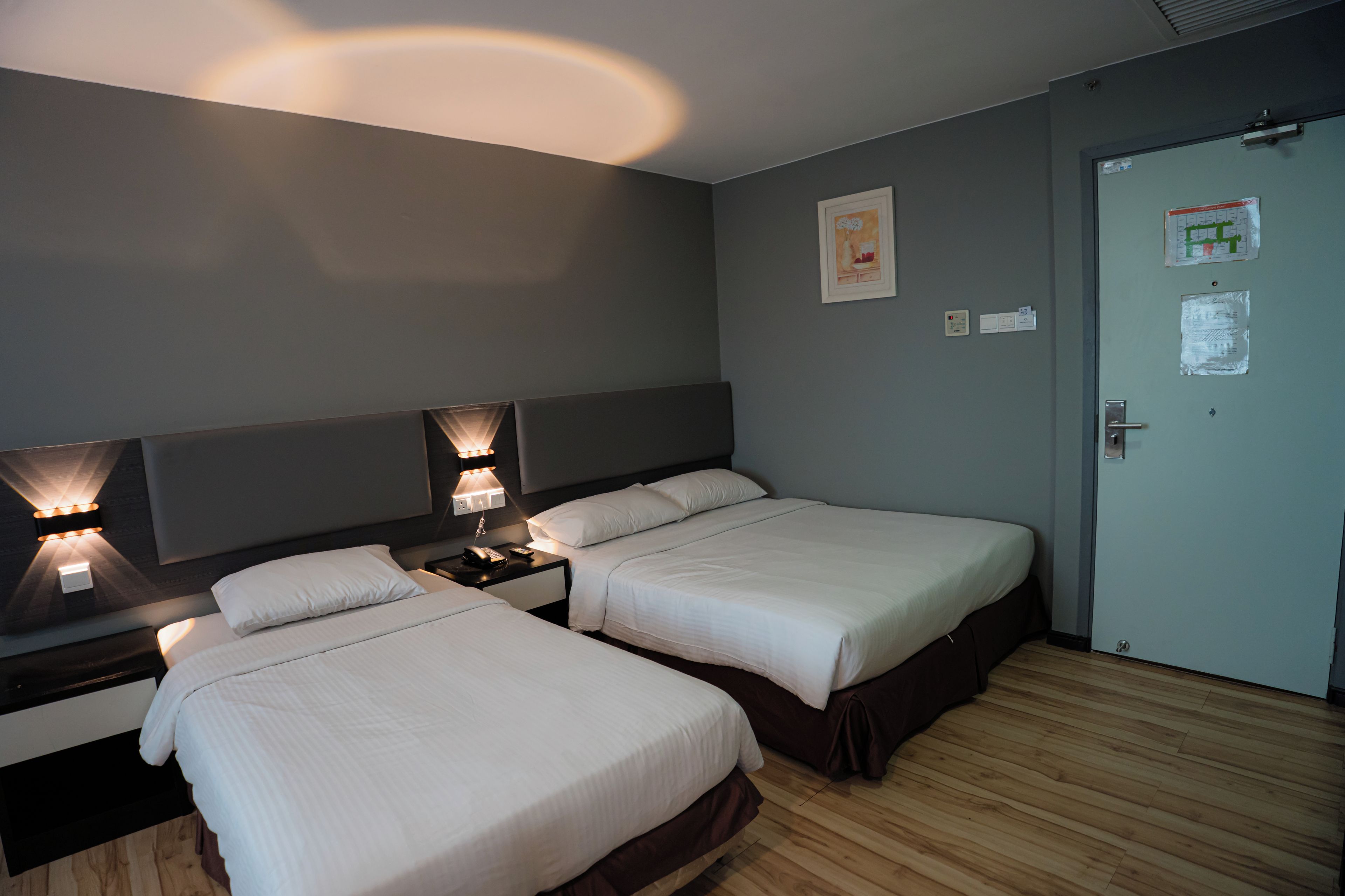 A hotel room with two beds, a nightstand with a phone, wall-mounted lights, and a door with a notice.