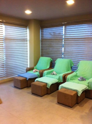 Una habitación con cuatro sillones de masaje verdes, cada uno con reposapiés, ubicados frente a amplias ventanas con persianas.