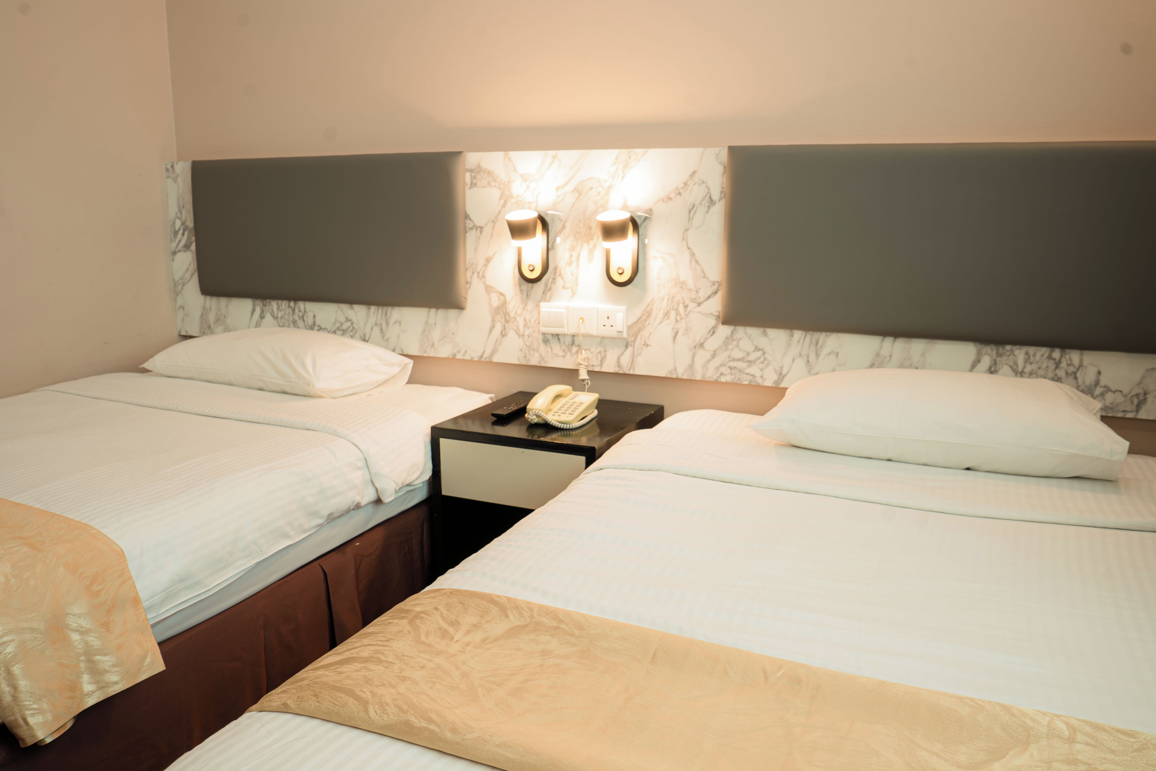 A hotel room with two beds, a nightstand with a telephone, and wall-mounted lighting.