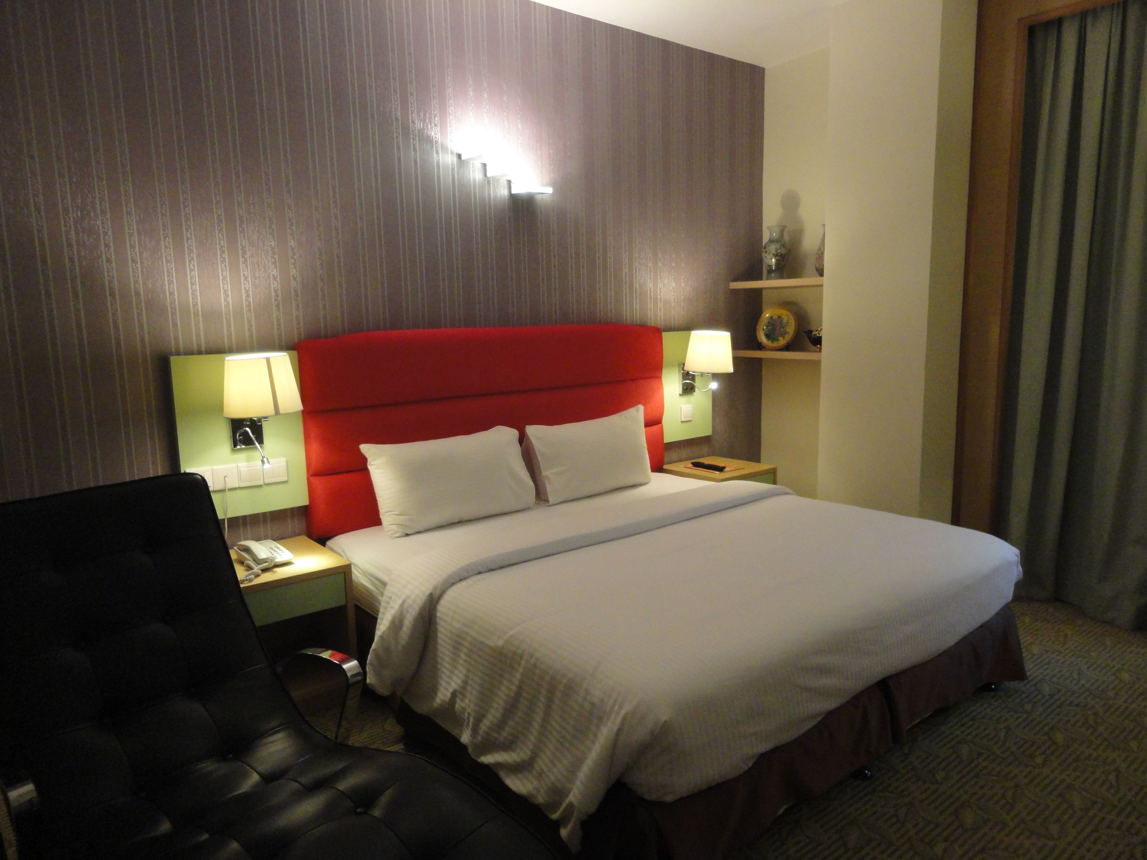 Ein Hotelzimmer mit einem großen Bett, einem Schreibtisch mit Stuhl, einem Nachttisch mit Lampe sowie einem Wandregal mit Dekoration.