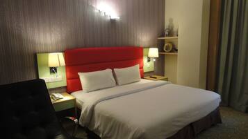 Ein Hotelzimmer mit einem großen Bett, einem Schreibtisch mit Stuhl, einem Nachttisch mit Lampe sowie einem Wandregal mit Dekoration.