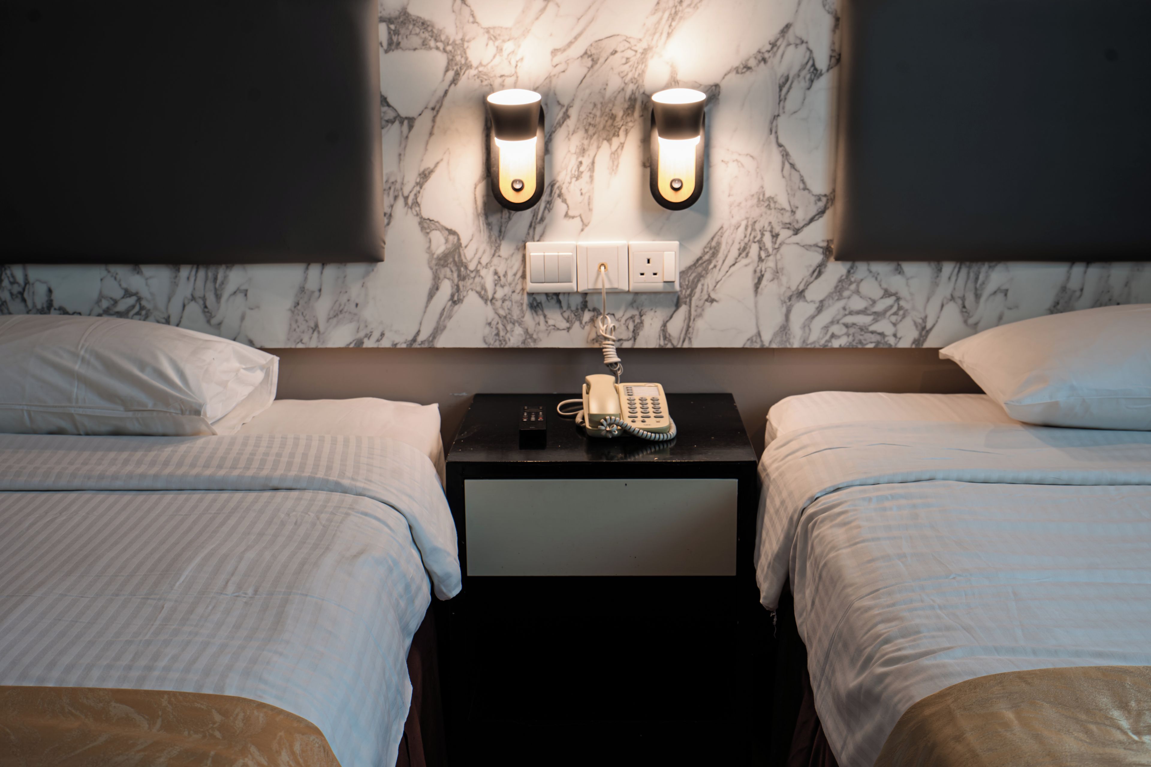 A hotel room with two beds, a nightstand with a telephone, and wall-mounted light fixtures.