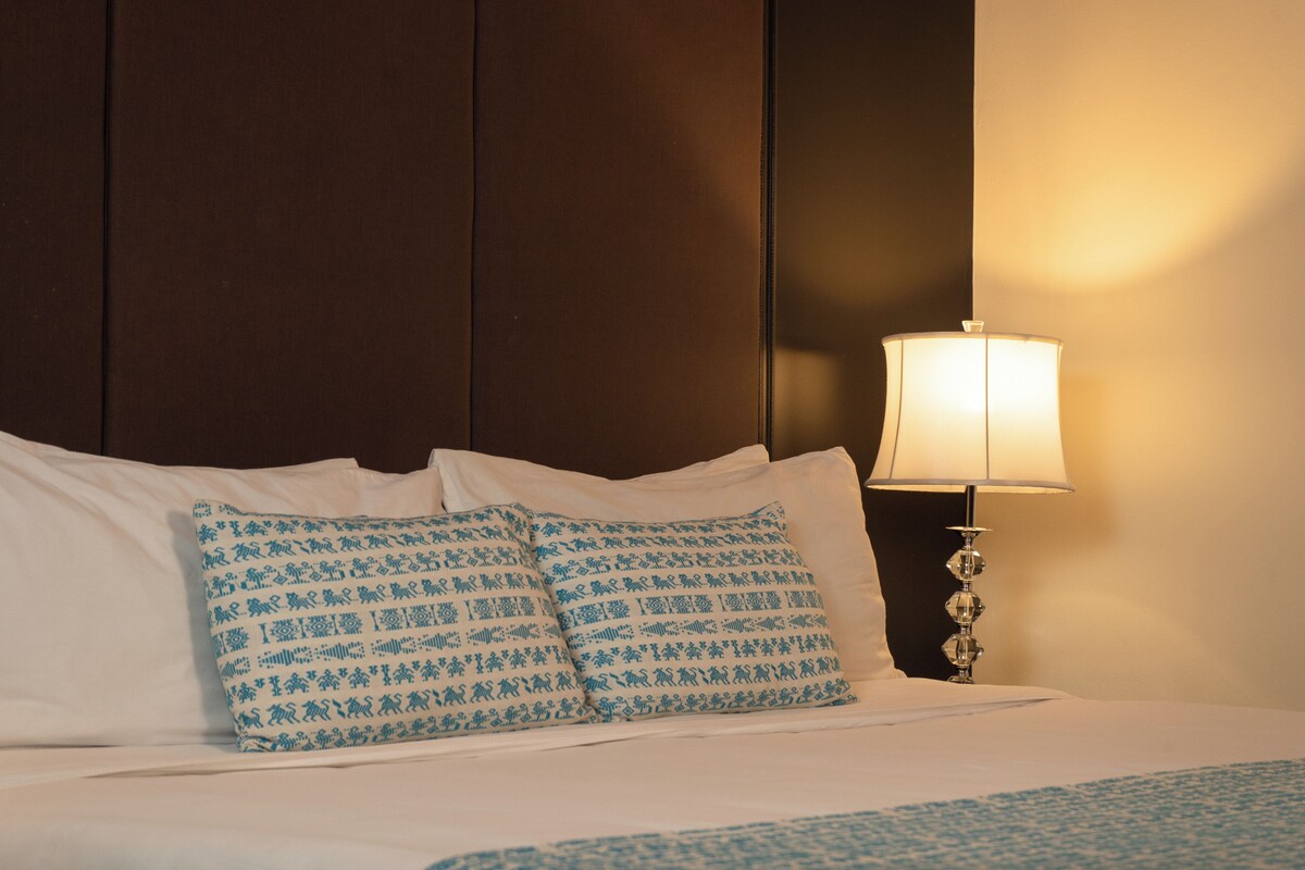 A neatly made bed with white sheets and two patterned pillows, a bedside lamp, and a dark wooden headboard.
