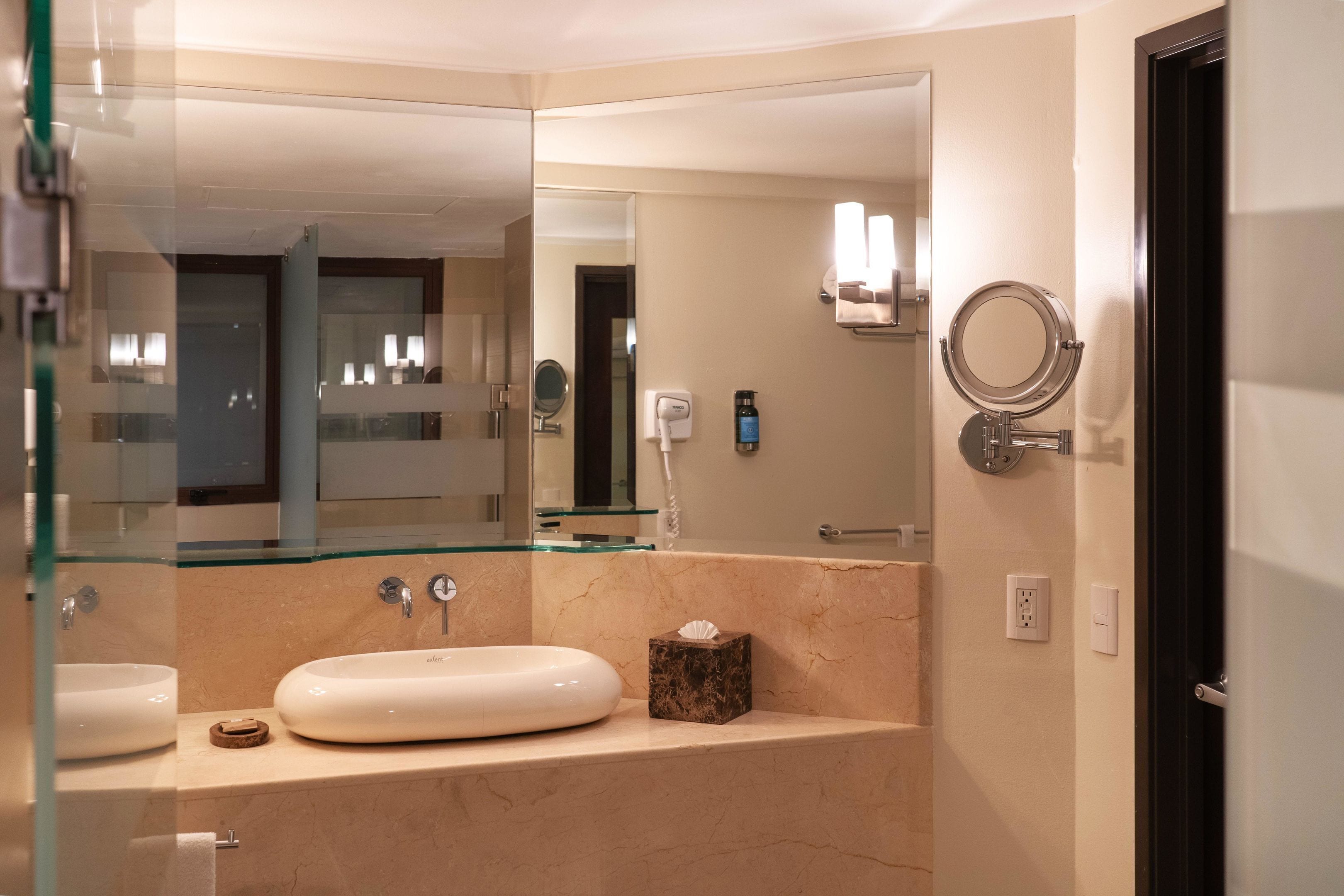 A modern bathroom with a glass-enclosed sink, a marble countertop, a large mirror, and a round mirror on the wall.