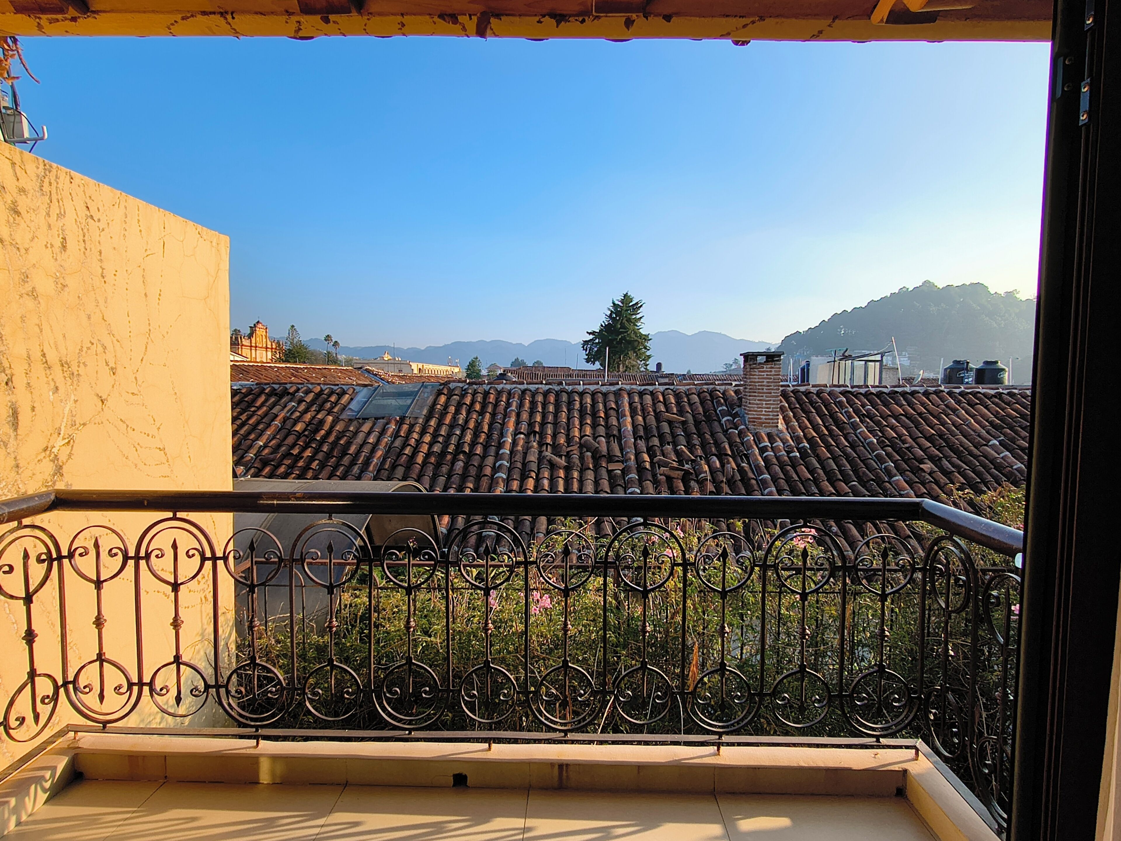 Un balcón con barandilla de hierro forjado, techo de tejas y vista a un paisaje urbano con edificios y montañas a lo lejos.