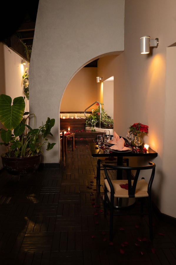 An outdoor dining area with a table set for two, surrounded by potted plants and candles.