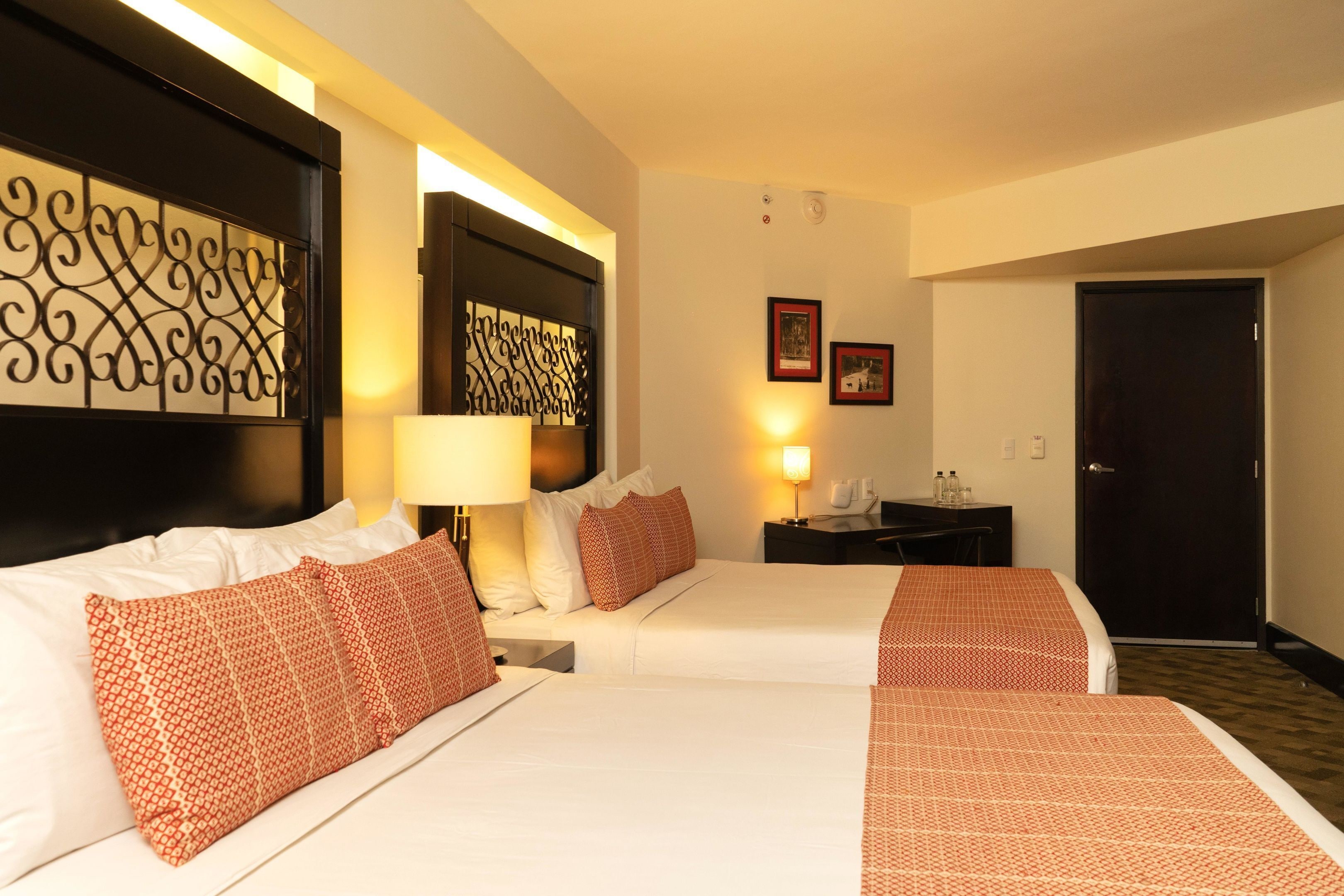 A hotel room with two beds, a nightstand, a lamp, framed pictures on the wall, and a door.