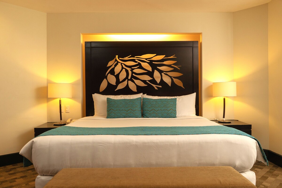 A hotel room with a large bed, two bedside lamps, a decorative wall panel, and a brown bench.