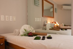 A spa room with two beds, a mirror, a wall-mounted air conditioner, and a wooden nightstand with a lamp and flowers.