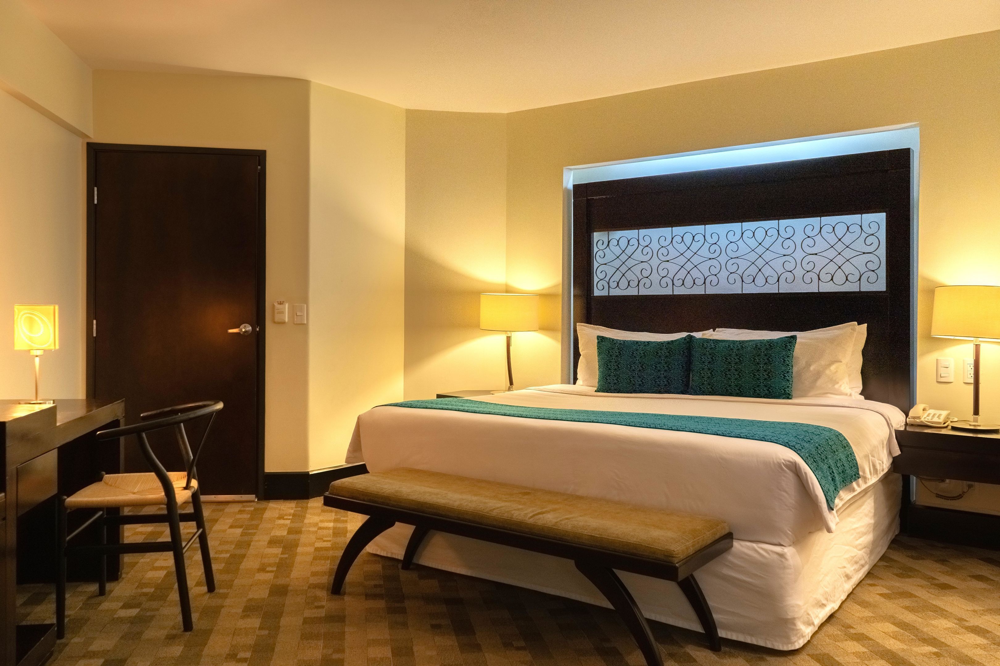 Una habitación de hotel con una cama, un escritorio, una silla, un banco y dos lámparas de noche.