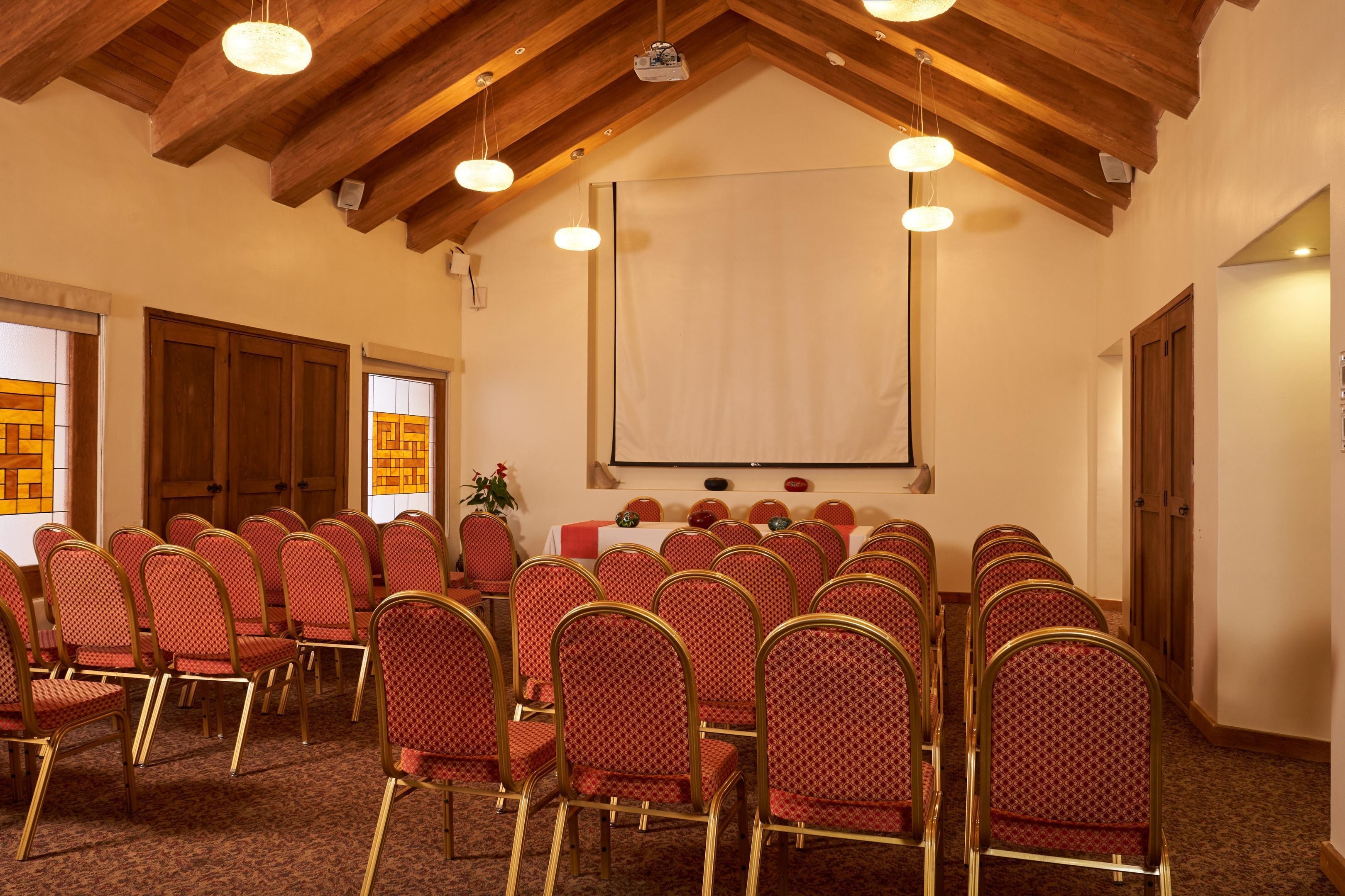 Une salle de conférence avec des rangées de chaises rouges, un écran de projection et des poutres en bois au plafond.