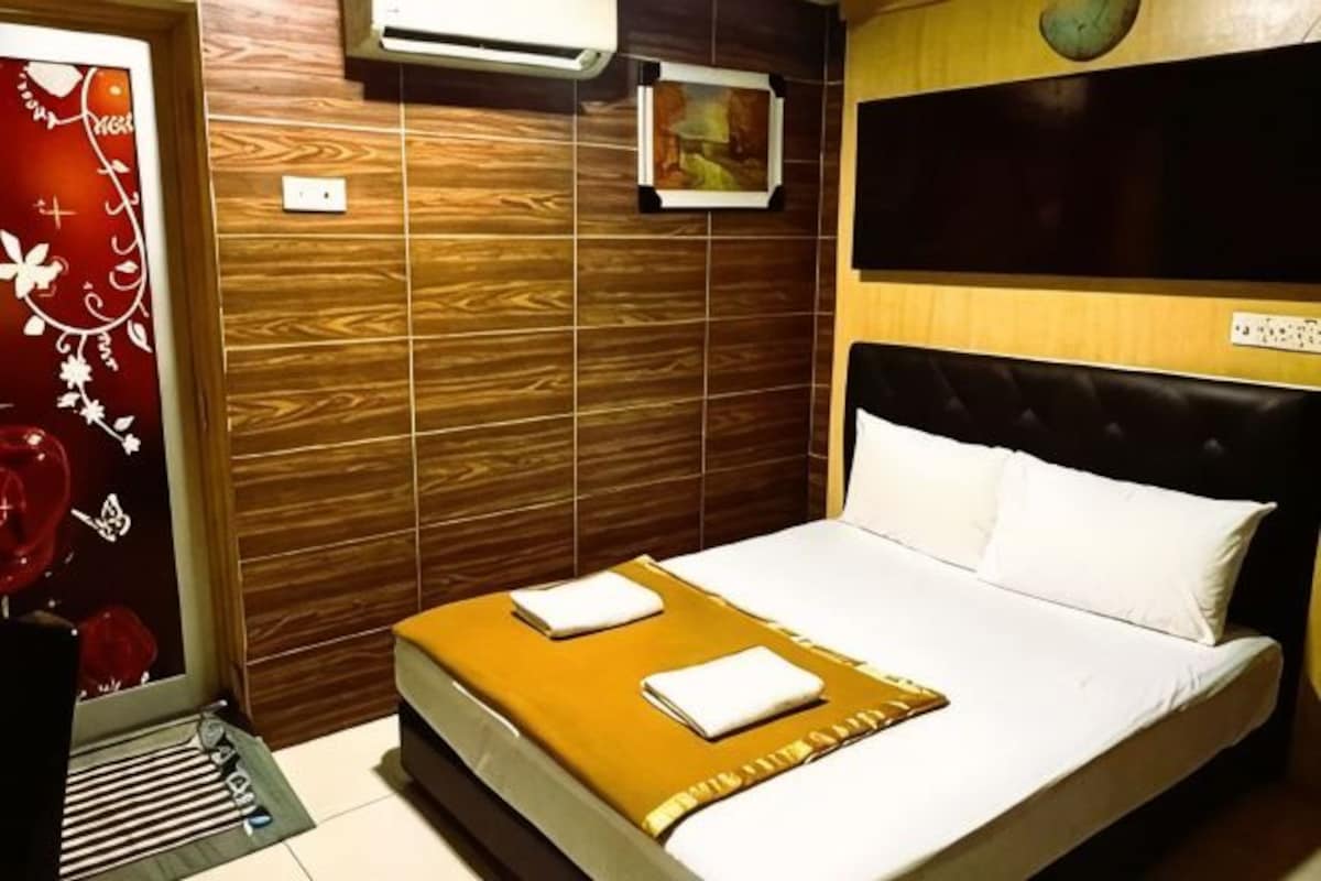 A hotel room with a bed, a wooden headboard, a wall-mounted air conditioner, and a small picture.