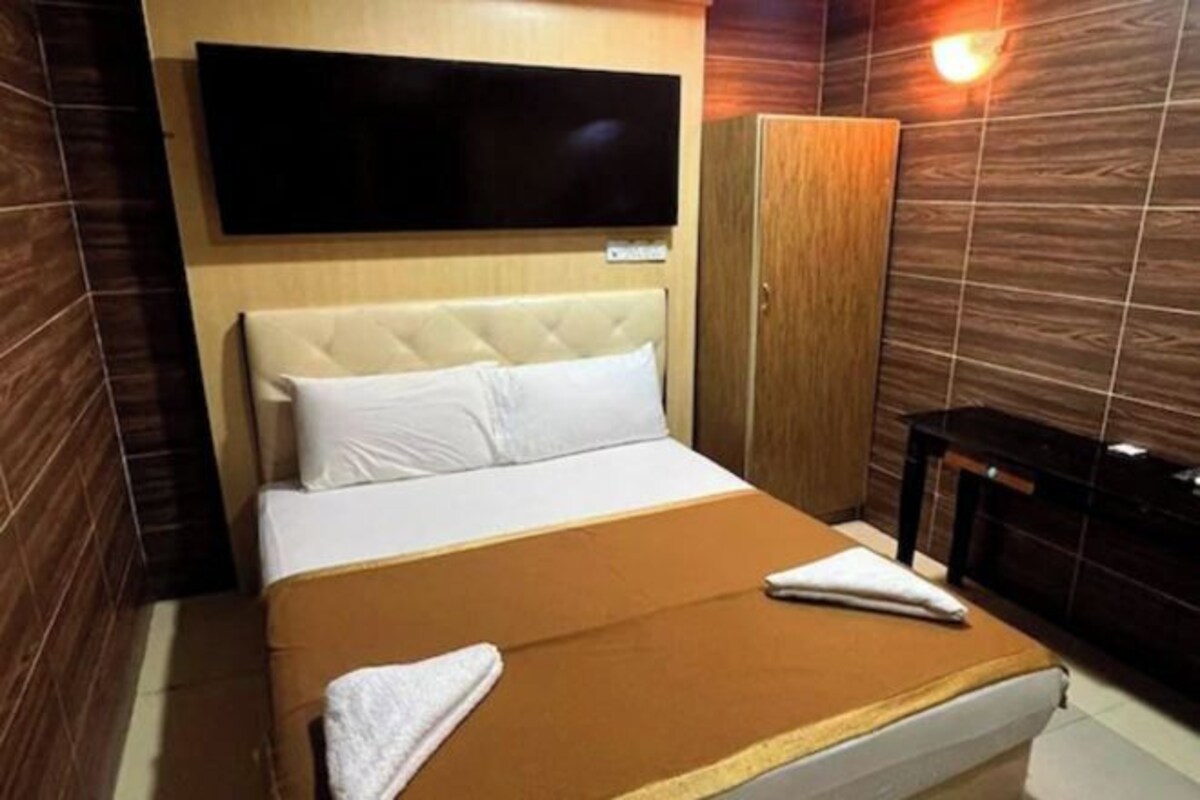 A hotel room with a bed, a television mounted on the wall, a wooden wardrobe, and a small table.