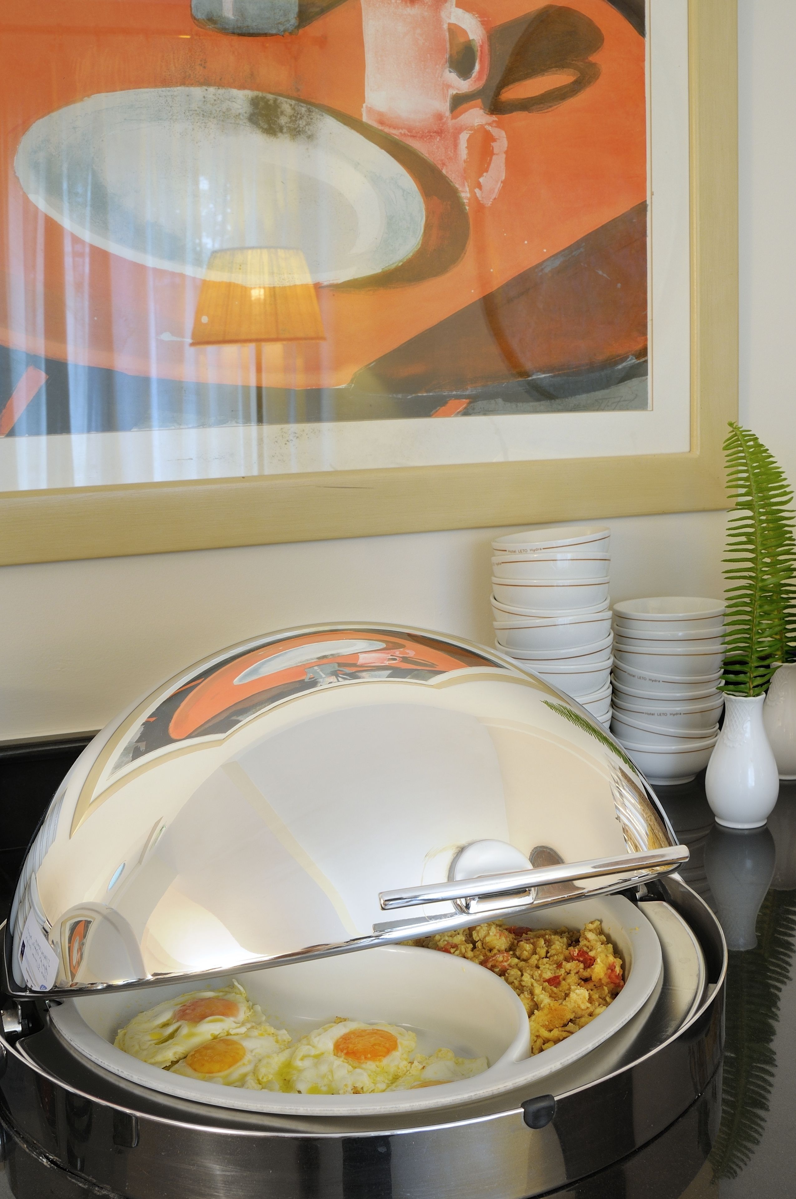 A buffet setup with a covered serving dish containing scrambled eggs and a side dish, a stack of white plates, and a potted fern.