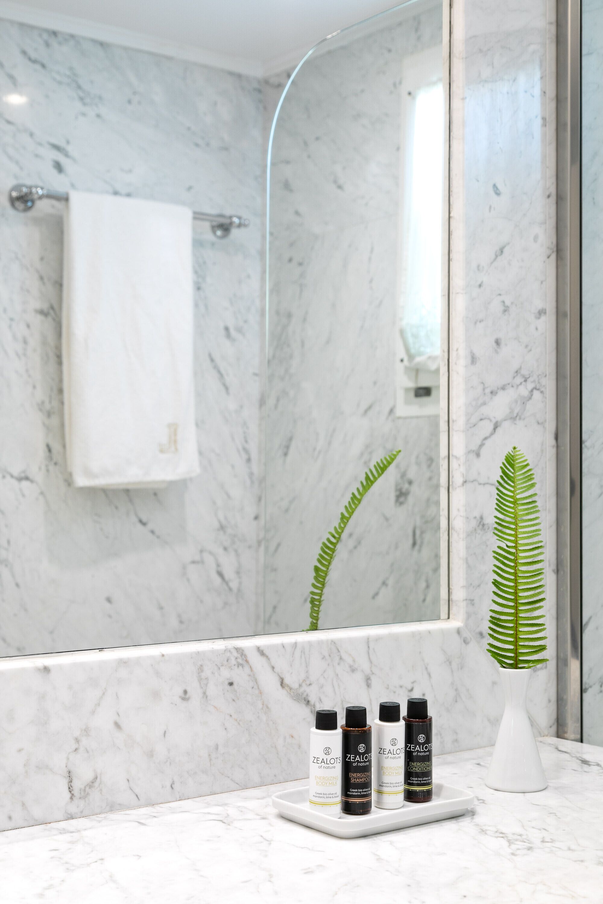 A bathroom with a marble wall, a white towel, a mirror, and three bottles of Zealot products on a tray.