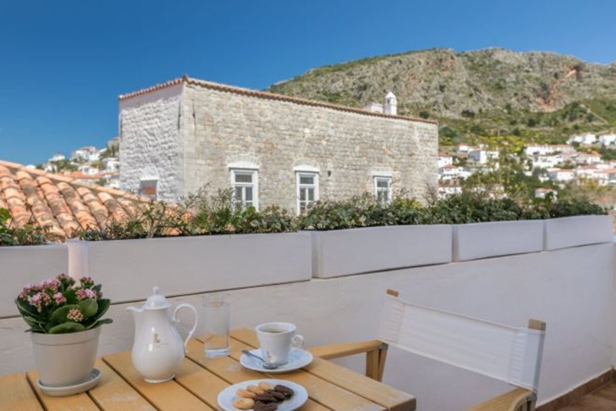 A terrace with a table set for two, a white pitcher, a cup, and a plate of cookies. A white wall and a stone building with a tiled roof are in the background.