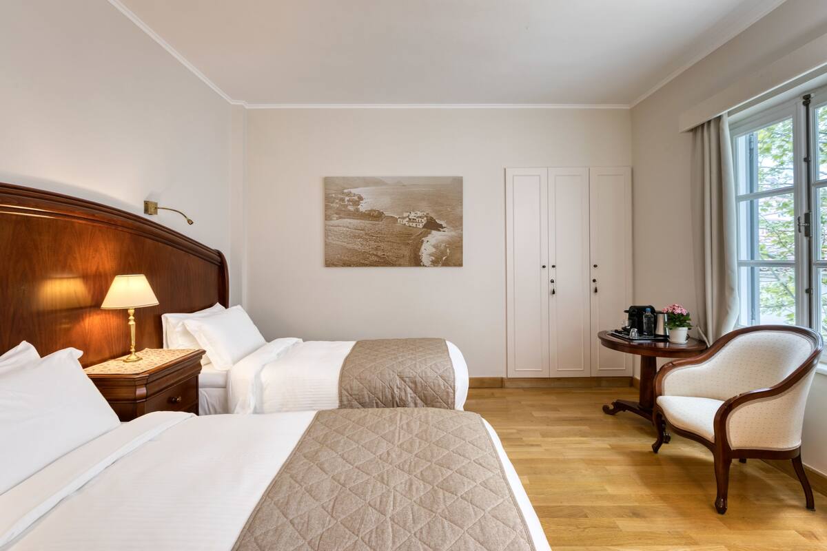 Habitación de hotel con dos camas, cabecero de madera, mesita de noche con lámpara, pequeña mesa redonda con cafetera, una silla y un cuadro en la pared.